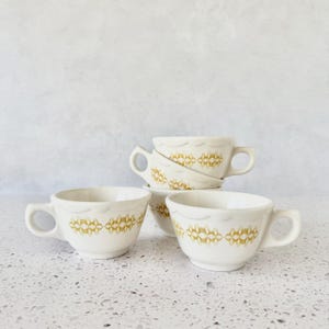 May include: Four white ceramic teacups with a gold geometric pattern and scalloped rims. The cups have a handle and are stacked and arranged on a white surface. The cups are vintage and have a mid-century modern design.