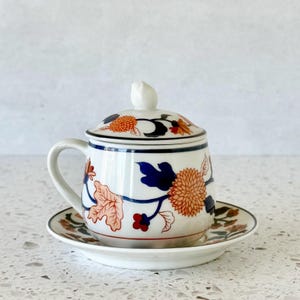 May include: A white ceramic teacup with a matching lid and saucer. The cup and lid feature a floral design in orange, blue, and red. The cup has a handle and sits on a small, round saucer. The set is designed for tea.