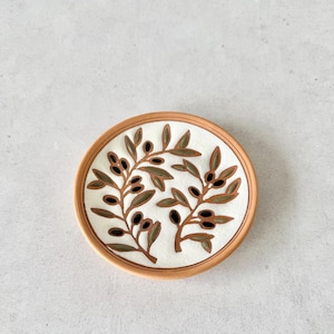 May include: A round ceramic dish with a terracotta-colored rim and a white center. The surface features a hand-painted design of two olive branches with green leaves and dark brown olives, arranged symmetrically across the circular plate base.