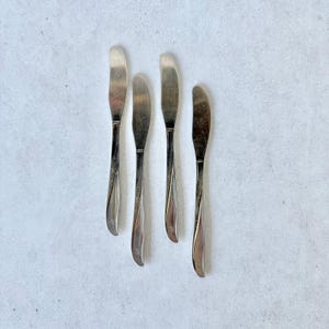 May include: Four silver-toned metal knives arranged vertically. Each knife features a smooth, rounded handle with a subtle, etched starburst pattern near the base of the handle. The blades are plain, polished, and have a rounded tip design.