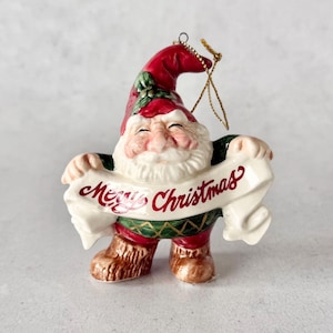May include: A ceramic Christmas ornament shaped like a gnome with a red hat, white beard, and green patterned tunic. The gnome holds a white banner with the red text Merry Christmas. A gold string is attached to the top of the pointed red hat.