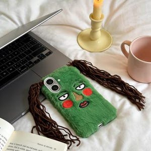 May include: A green phone case with a cartoon face design, featuring brown hair extensions and red blush. The case is placed on a white surface next to a laptop, a candle, and a pink mug.