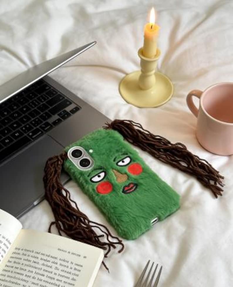 May include: A green phone case with a cartoon face design, featuring brown hair extensions and red blush. The case is placed on a white surface next to a laptop, a candle, and a pink mug.
