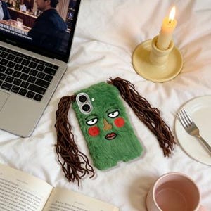 May include: A green, furry phone case with an embroidered face and brown hair extensions. The case features a cartoon face with red cheeks, a brown nose, and a red mouth. The phone case is lying on a white bed.