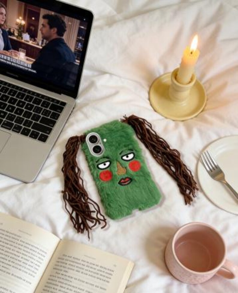 May include: A green, furry phone case with an embroidered face and brown hair extensions. The case features a cartoon face with red cheeks, a brown nose, and a red mouth. The phone case is lying on a white bed.