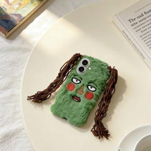 May include: A green, fuzzy phone case featuring an embroidered face with red cheeks, a brown nose, and a mouth. The case has brown, braided hair extensions. The phone case is on a cream-colored surface.