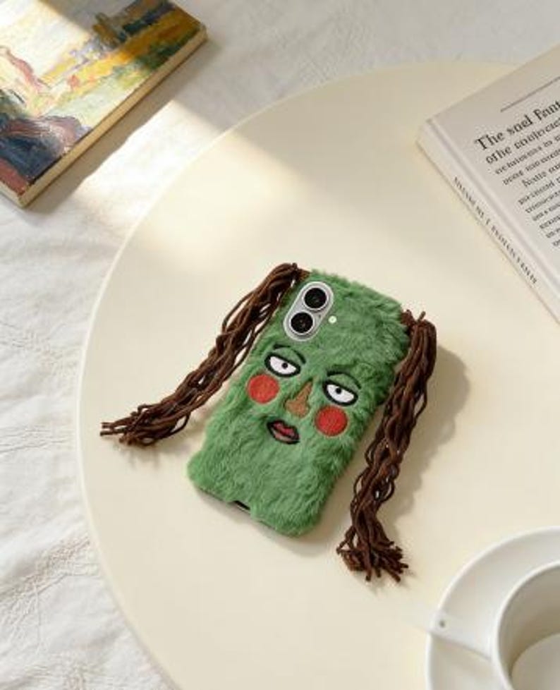 May include: A green, fuzzy phone case featuring an embroidered face with red cheeks, a brown nose, and a mouth. The case has brown, braided hair extensions. The phone case is on a cream-colored surface.