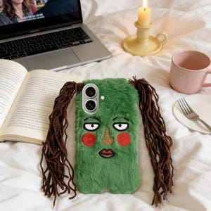 May include: A green, furry phone case with an embroidered face and long brown hair extensions. The case features a cartoon face with red cheeks, a gold nose, and a red lip. The phone case is lying on a white bedspread.