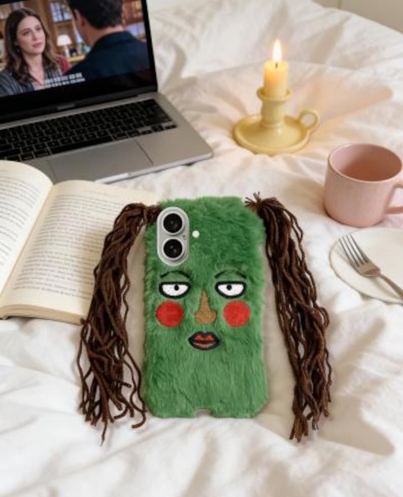 May include: A green, furry phone case with an embroidered face and long brown hair extensions. The case features a cartoon face with red cheeks, a gold nose, and a red lip. The phone case is lying on a white bedspread.