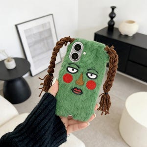 May include: A green, fuzzy phone case with an embroidered face design featuring red cheeks, a brown nose, and a red lip. The case has brown braided hair extensions attached to the top. The phone case is held in a person's hand.