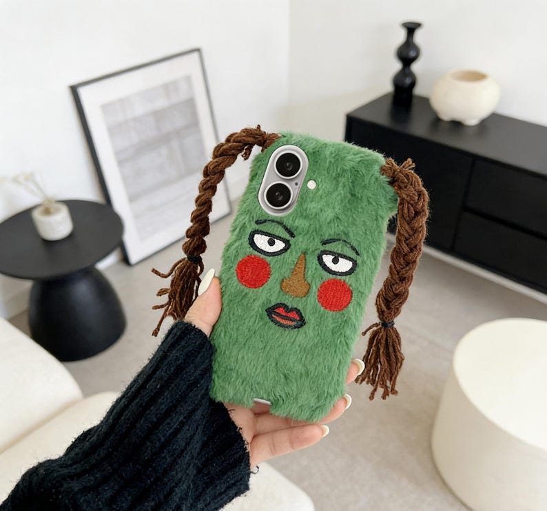 May include: A green, fuzzy phone case with an embroidered face design featuring red cheeks, a brown nose, and a red lip. The case has brown braided hair extensions attached to the top. The phone case is held in a person's hand.