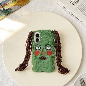 May include: A green, furry phone case with an embroidered face featuring a brown nose, red cheeks, and a dark red mouth. The case has long, brown, braided hair on each side. The phone case is on a cream-colored plate.