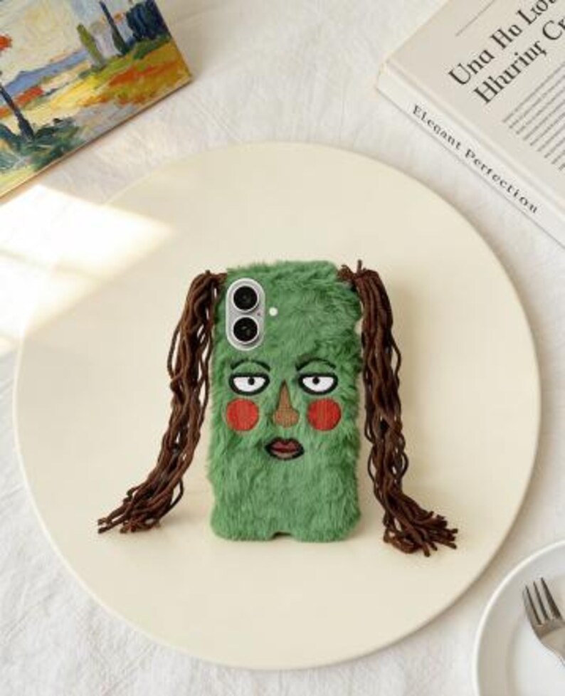 May include: A green, furry phone case with an embroidered face featuring a brown nose, red cheeks, and a dark red mouth. The case has long, brown, braided hair on each side. The phone case is on a cream-colored plate.