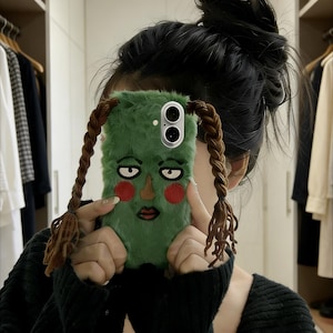 May include: A green, fuzzy phone case featuring a cartoon face with red cheeks, a brown mouth, and brown braids. The case has a dual-lens camera cutout and is held in front of a person.
