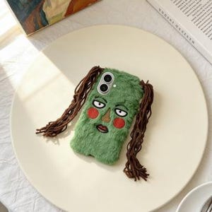 May include: A green, fuzzy phone case featuring an embroidered face with red cheeks, a brown nose, and a downturned mouth. The case has long, brown, braided hair-like strands. The phone case is on a cream-colored plate.