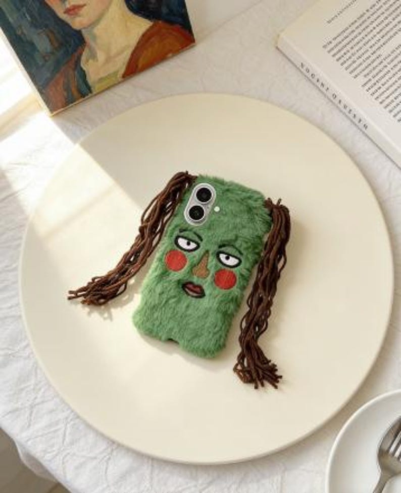 May include: A green, fuzzy phone case featuring an embroidered face with red cheeks, a brown nose, and a downturned mouth. The case has long, brown, braided hair-like strands. The phone case is on a cream-colored plate.