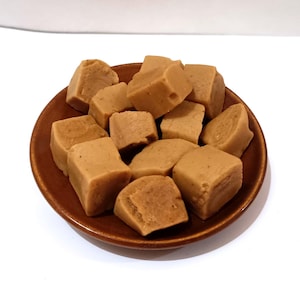 May include: A brown ceramic plate filled with light brown, square-shaped fudge pieces. The fudge has a slightly rough texture and is arranged in a pile. The plate is round with a dark brown rim, set against a white background.