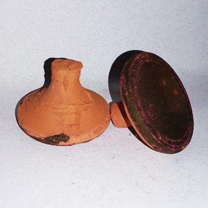 Op de afbeelding: Twee terracotta-klei flessenstoppers. Eén heeft een afgeronde basis en een korte, cilindrische hals. De andere is een platte, cirkelvormige schijf met een klein handvat. De schijf heeft een donkere, gevlekte uitstraling.