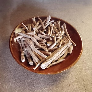 May include: A brown ceramic bowl filled with dried, light brown, and beige root pieces. The roots are of varying lengths and thicknesses, with a slightly rough texture. The bowl is round with a dark brown rim.