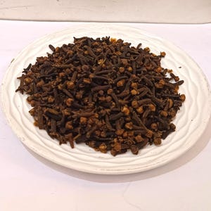 May include: A white, scalloped-edge plate filled with whole cloves. The cloves are a deep brown color, with small, round heads and slender stems. The plate is set against a white background.