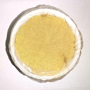 May include: A top-down view of a white bowl filled with a yellow powder. The bowl is lined with a clear plastic bag. The powder has a fine, granular texture and appears to be a light shade of yellow.
