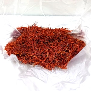 Puede incluir: Un montón de hebras de azafrán de color rojo anaranjado brillante, una especia culinaria, se encuentra dentro de una bolsa de plástico blanca translúcida. Las hebras están densamente empaquetadas, mostrando su delicada textura y rico color.