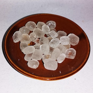 Peut inclure: Un ensemble de cristaux translucides de forme irrégulière, possiblement des pierres précieuses ou des minéraux, est disposé dans une petite coupelle ronde en céramique marron. Les cristaux ont un aspect légèrement givré.