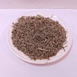 May include: A white ceramic plate piled high with dried, brown herbs. The herbs are thin and needle-like, filling the plate. The plate is round with a raised edge, set against a pale background. The herbs appear to be dried.