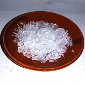 Puede incluir: Un plato pequeño y redondo de cerámica marrón lleno de un montón de estructuras cristalinas transparentes. Los cristales tienen una variedad de formas y tamaños, algunos parecen fragmentos y otros más en bloque. El plato está sobre un fondo blanco.