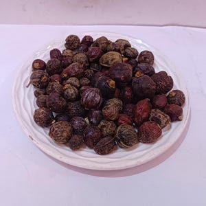 以下が含まれることがあります： 白のセラミックプレートに、乾燥した濃い茶色と赤褐色のベリーが盛り付けられています。 ベリーは質感のある表面を持ち、視覚的に面白い配置になるように集まっています。