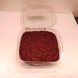 Può includere: Un contenitore di plastica trasparente, parzialmente aperto, riempito con materiale sminuzzato marrone-rossastro. Il contenitore è su una superficie bianca, con il coperchio leggermente socchiuso. Il materiale sembra essere distribuito uniformemente.