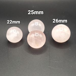May include: Four rose quartz spheres of varying sizes are displayed against a dark background. The spheres are labeled with their diameters: 22mm, 25mm, and 26mm. The spheres have a translucent, pinkish hue.