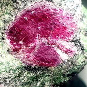 May include: A close-up of a rough, green gemstone with a large, bright pink ruby embedded in the center.