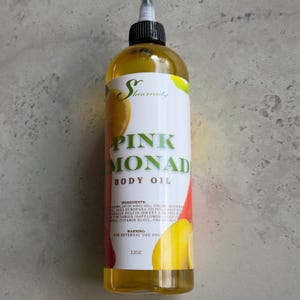 May include: A clear glass bottle with a yellow liquid inside. The bottle has a white label with the text "Shenrenity" in a cursive font. The label also has the text "PINK MONAD BODY OIL" in black text. The ingredients and warning are listed on the label. The bottle is sitting on a gray surface.