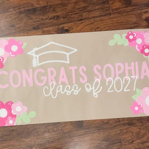May include: A graduation banner with the text "Congrats Sophia class of 2027" in pink and white. The banner is decorated with pink and white flowers and a graduation cap. The banner is on a brown background.