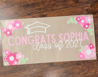 May include: A graduation banner with the text "Congrats Sophia class of 2027" in pink and white. The banner is decorated with pink and white flowers and a graduation cap. The banner is on a brown background.