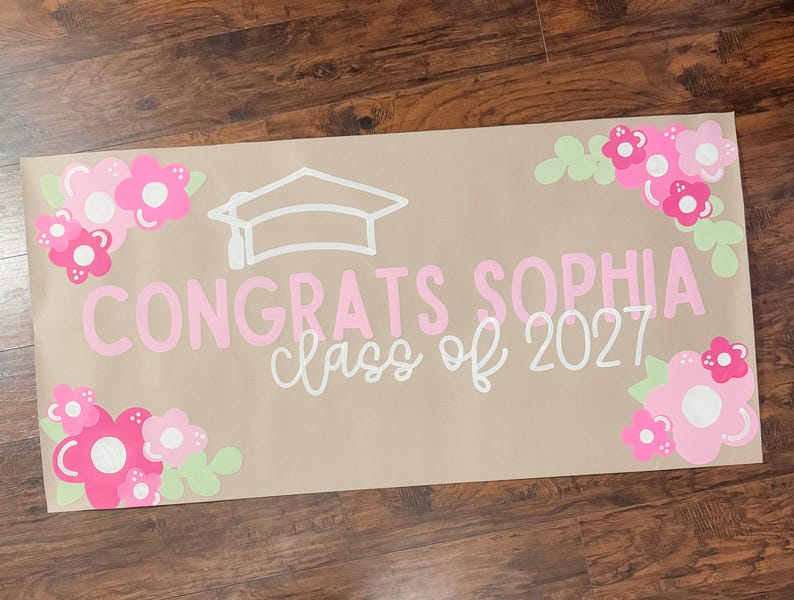 May include: A graduation banner with the text "Congrats Sophia class of 2027" in pink and white. The banner is decorated with pink and white flowers and a graduation cap. The banner is on a brown background.