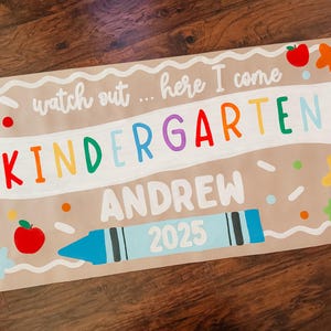 May include: A rectangular sign on a tan background featuring the text "watch out... here I come" in white, "KINDERGARTEN" in multicolored letters, "ANDREW" in white, and "2025" inside a light blue crayon graphic, decorated with red apples and stars.