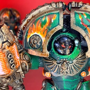 May include: A miniature figurine of a warrior in green and gold power armor with flame patterns on the shoulder pauldrons. The figure features a black helmet with glowing red eyes and holds a large weapon with bright orange glowing coils.