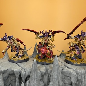 May include: Three miniature figurines of alien creatures with red blades and purple accents. The figures are posed on a gray, rocky terrain, with a yellow background. The creatures have a skeletal appearance and are painted with detailed features.