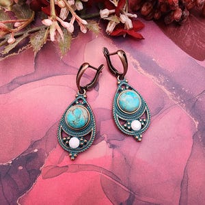 May include: A pair of turquoise and bronze-coloured earrings. Each earring features a teardrop shape with a turquoise stone centre, surrounded by a bronze-coloured frame. The earrings have a decorative design with white accents and a hook closure.