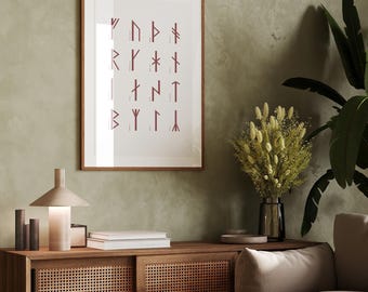May include: A framed print of the runic alphabet, with each rune in a dark red color, hangs on a light green wall. A vase with dried white flowers sits on a wooden table with a lamp and books. A light brown couch with a white throw pillow is in the background.