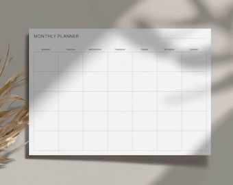 May include: A white monthly planner with a grid layout for scheduling. The planner has the words "MONTHLY PLANNER" at the top, with days of the week listed across the top row. The planner is on a light gray surface with shadows.