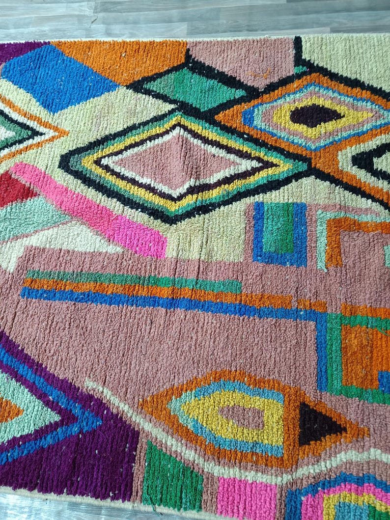 May include: A colorful, geometric rug with a textured surface. The rug features a variety of shapes, including diamonds, squares, and triangles, in shades of pink, blue, green, orange, and yellow. The rug's design is a mix of abstract and traditional patterns.