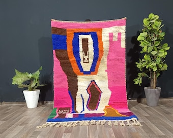 May include: A vibrant, hand-woven rug with a bold, abstract design. The rug features a striking combination of colors, including bright pink, blue, orange, brown, and white. The rug has a fringed edge and is displayed against a dark background.