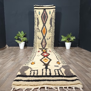 Puede incluir: Alfombra de pasillo color crema con patrones geométricos en negro, gris, naranja y rojo. La alfombra presenta formas de diamantes y animales abstractos, con un borde negro y flecos. Un revestimiento de suelo decorativo.
