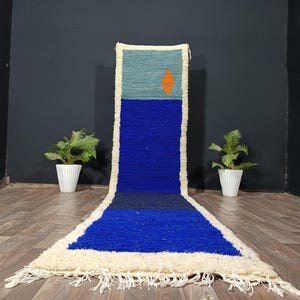 Puede incluir: Alfombra rectangular hecha a mano con una combinación de colores azul y crema. La alfombra presenta un campo azul con una sección superior turquesa y un rombo naranja. La alfombra tiene un borde color crema con flecos.