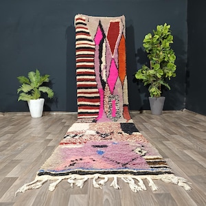 Puede incluir: Una alfombra de pasillo larga con una mezcla de patrones geométricos y colores que incluyen rosa intenso, naranja, rojo, negro y crema. El diseño presenta rayas horizontales y formas de diamante, terminado con flecos color crema en el borde.