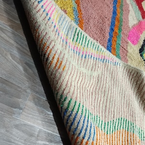 May include: A colorful, hand-woven rug with a geometric design. The rug features a cream-colored border with vertical stripes in orange, green, and blue. The central design includes pink, blue, yellow, and black shapes on a pink background.