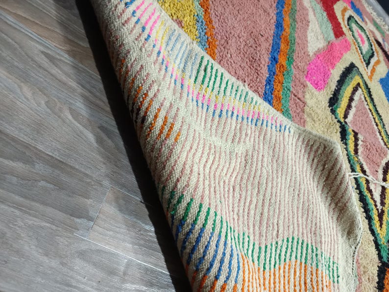 May include: A colorful, hand-woven rug with a geometric design. The rug features a cream-colored border with vertical stripes in orange, green, and blue. The central design includes pink, blue, yellow, and black shapes on a pink background.